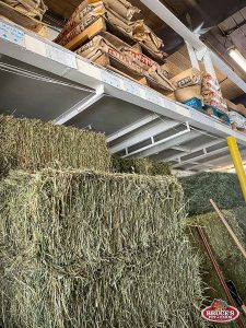 Bruce's Pet and Farm Supply storage area with stacked hay bales and bags of livestock feed on upper shelves. The area is stocked with essential feed and bedding supplies for farm animals, including high-quality hay for livestock nutrition. A convenient, organized setup designed to serve the needs of farmers and pet owners in the Maple Ridge area.