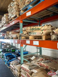 Bruce's Pet and Farm Supply storage racks filled with large bags of livestock feed, including Lifeline and Otter Co-op Feed brands. Shelves are stacked high with various types of animal feed, providing ample stock for local farmers and animal caretakers. The organized warehouse section caters to the nutritional needs of livestock and farm animals, ensuring quality feed options are readily available.